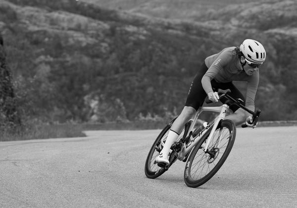 Black and white shot of Christian cycling round a road bend, mountainous landscape behind. 