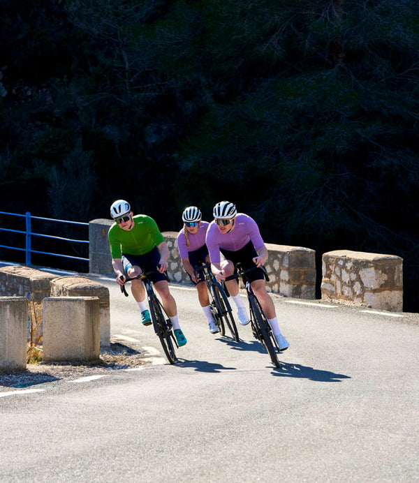 Three cyclists in purple or green jerseys riding towards round a bend.