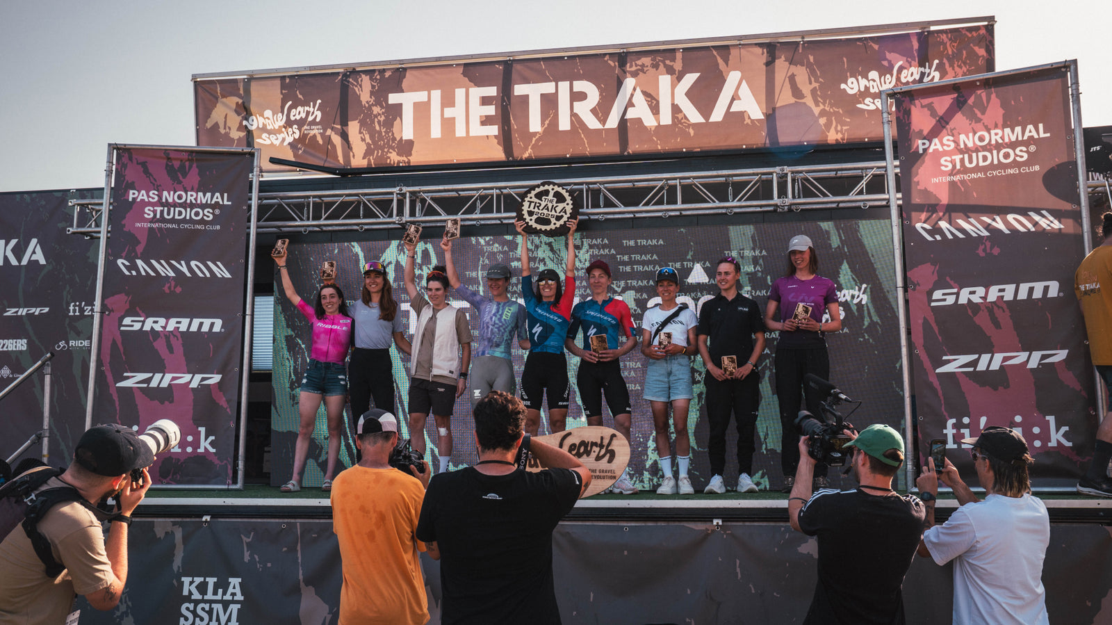 Group of cyclists on the Traka stage podium, surrounded by multiple sponsor logos in the background