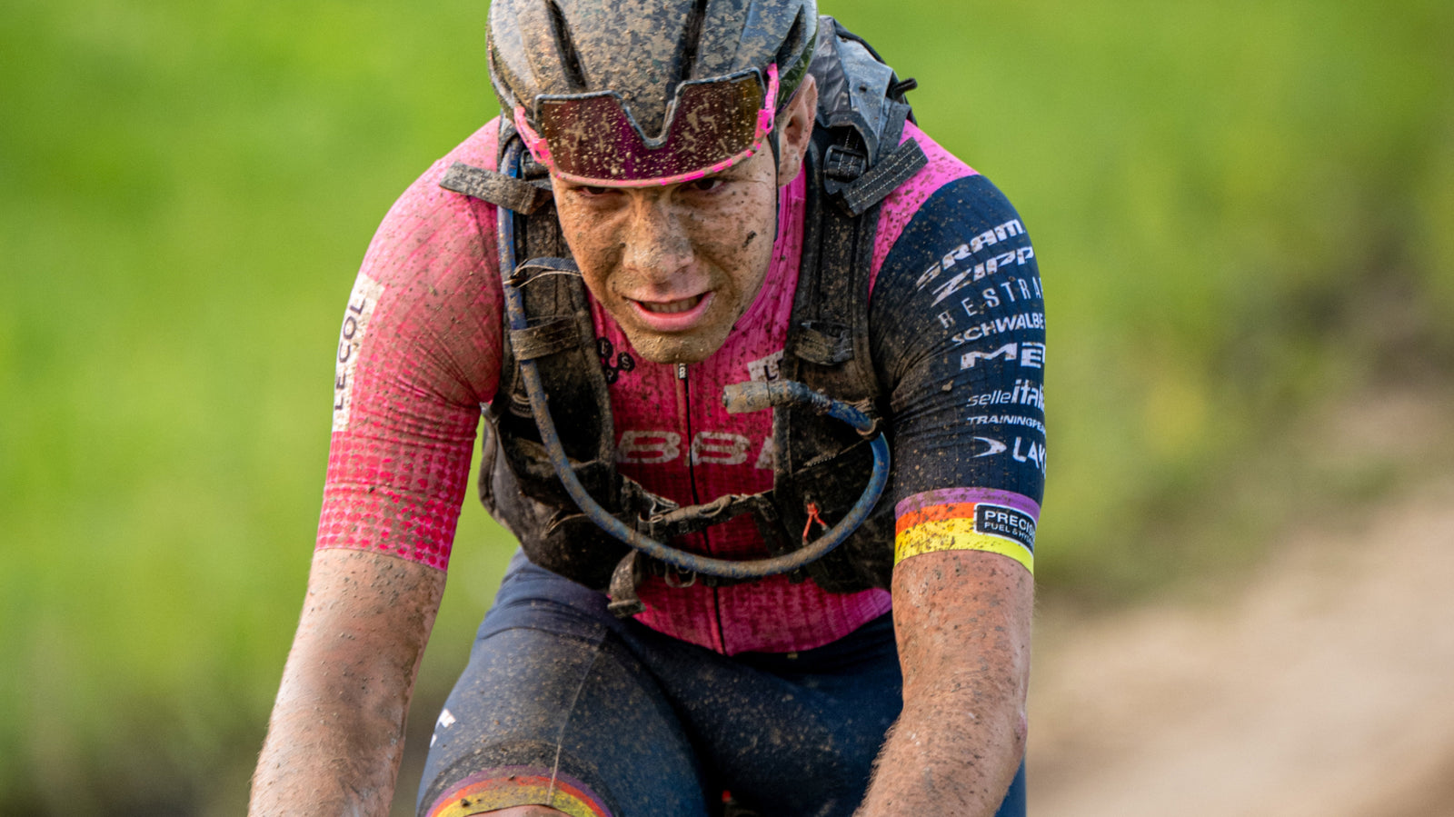 Cyclist in a racing jersey with mud on their face and body, riding on a gravel path.