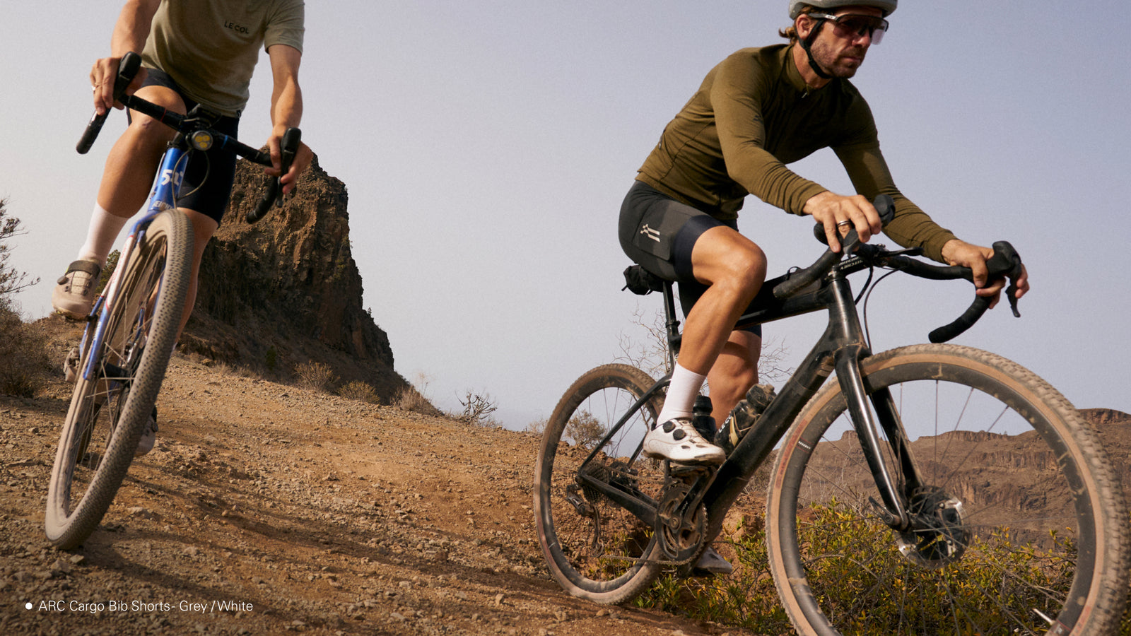 low-level shot of two male gravel riders riding downhill, rocky outcrop behind