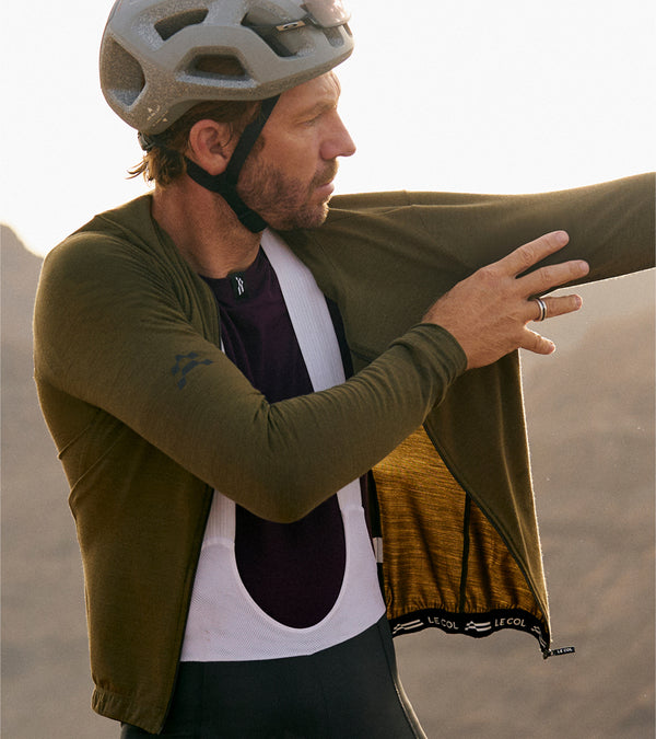 Male rider pulling on olive long sleeve jersey with grey base layer underneath.