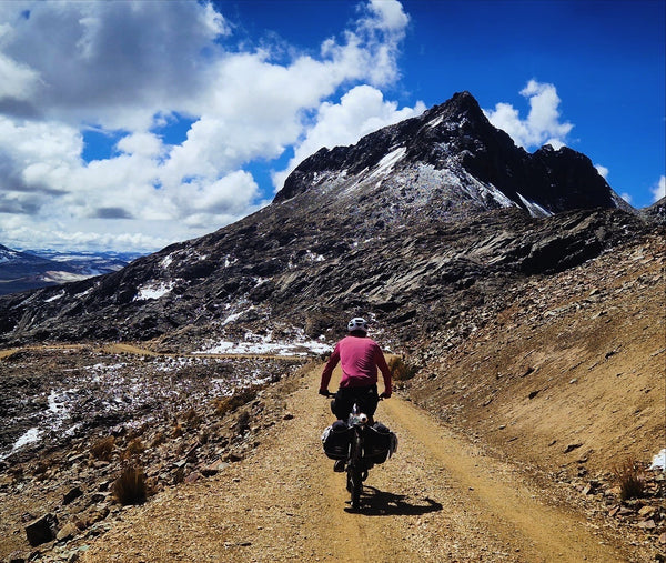 Angus cycling on mountain dirt track with peaks rising above with a bright blue sky.