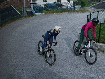 Two cyclists on a track, one in a blue outfit and helmet, the other in a pink outfit and helmet, with a residential area in the background.