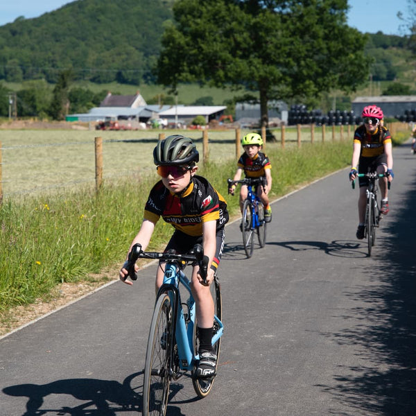 Two children and one adult cycling on a countryside path with a farm in the background.
