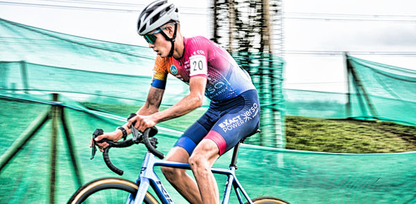 Young male cyclist pedalling hard in colourful kit, green track barriers behind.