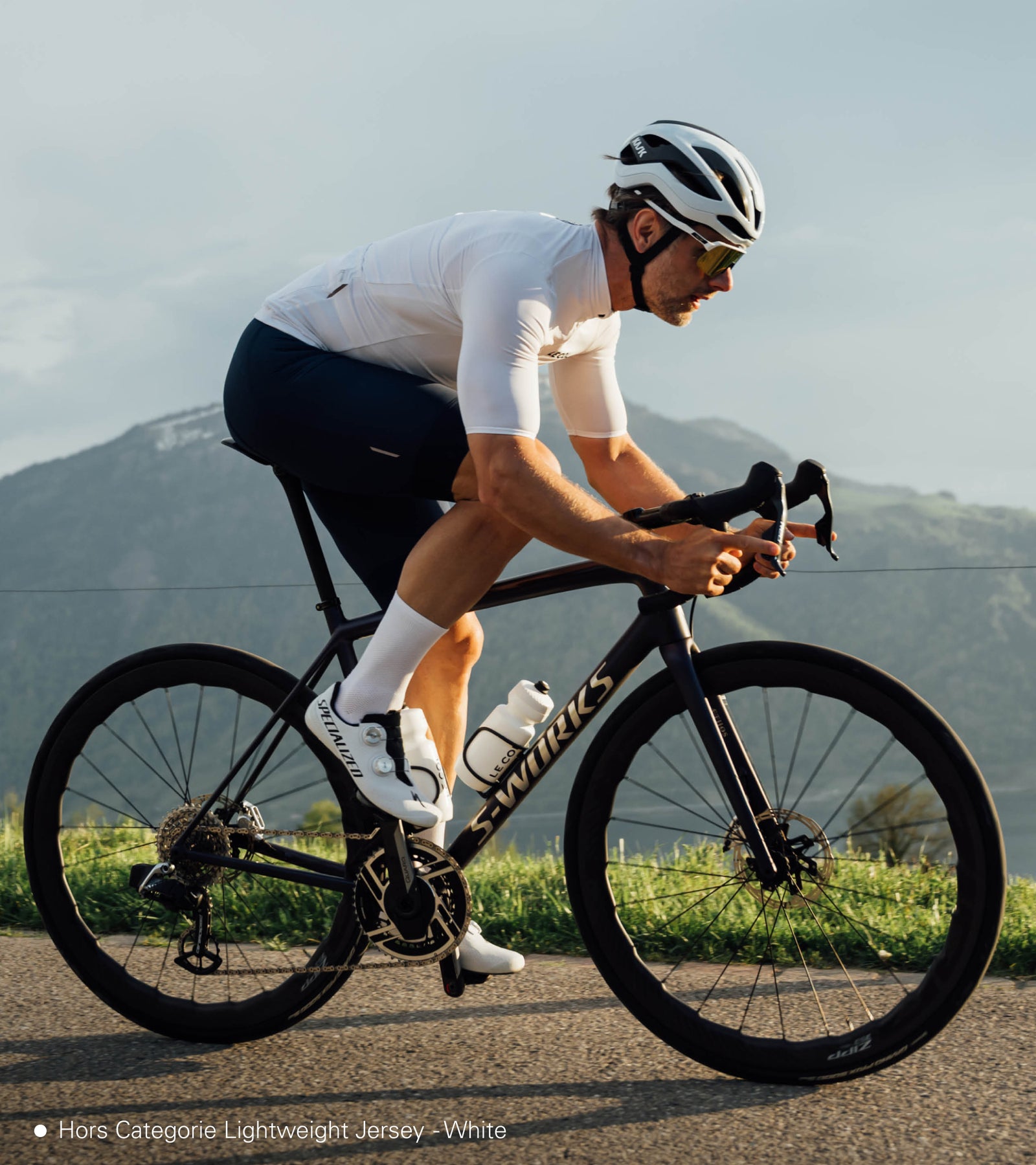 Male cyclist riding in a white jersey with mountains in the background.