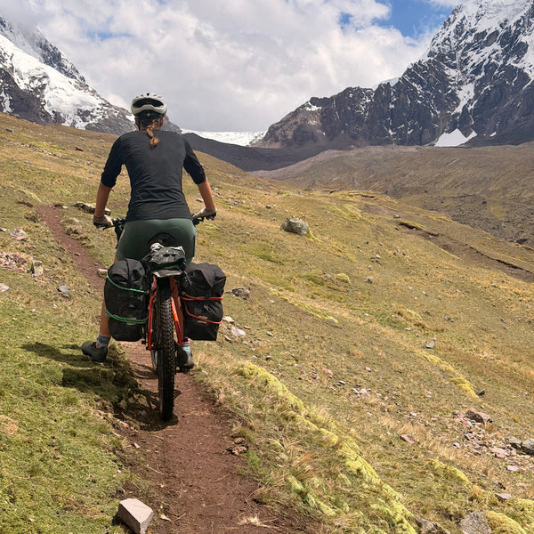 Angus' Wife cycling on dirt track on a grassy mountainside with snow capped peaks ahead.