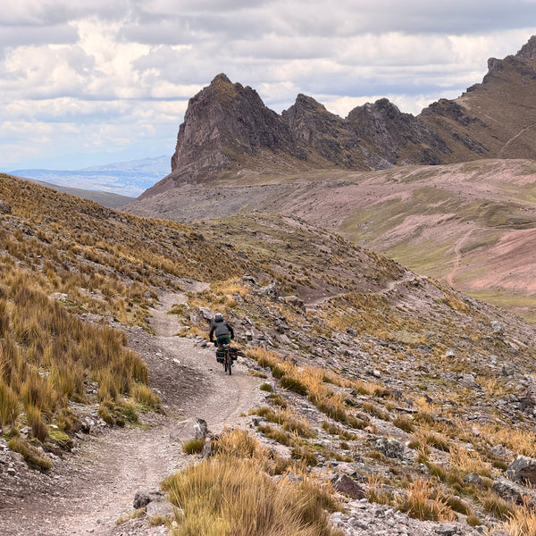Angus cycling in the distance on a gravely mountainside track with peaks behind.