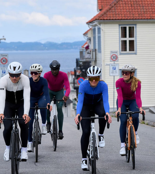 Five cyclists riding towards on a road with buildings and the sea behind.