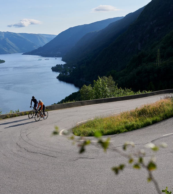Two cyclists on a hairpin bend road with a Fjord behind.