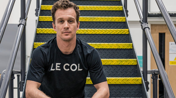 Colin in black Le Col T-shirt, sat on metal stairs.