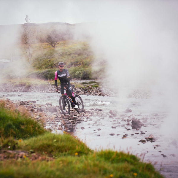 Male cyclist paused on his bike in a rocky stream with steam rising obscuring half the image. 