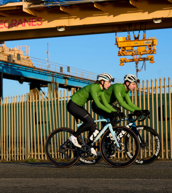 Two cyclists in green long sleeve jerseys riding along a fence in a dockyard setting.