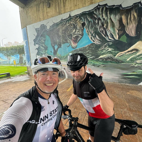 Bike selfie of Shawn and his friend in cycling kit in front of a t-rex mural.