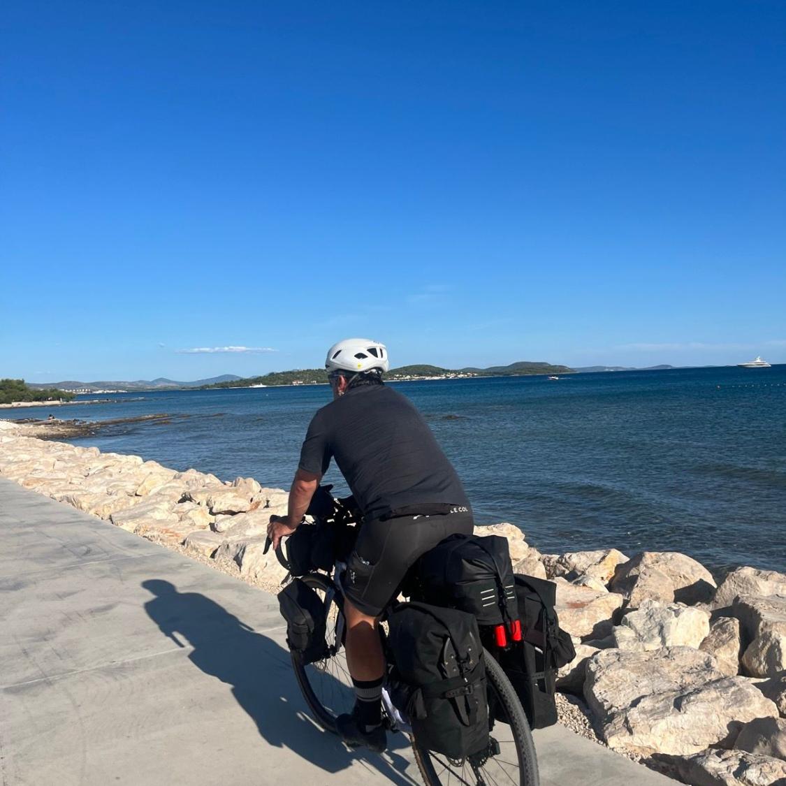 Person with a bicycle near a coastal area with clear blue sky and ocean.