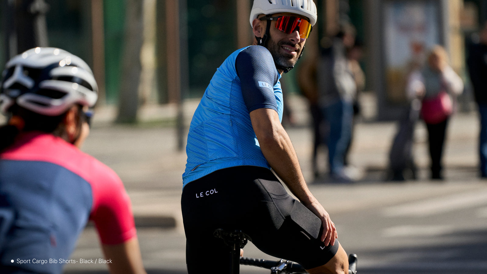 male cyclist in black bibshorts and blue jersey stood up on bike, half turned to face backwards