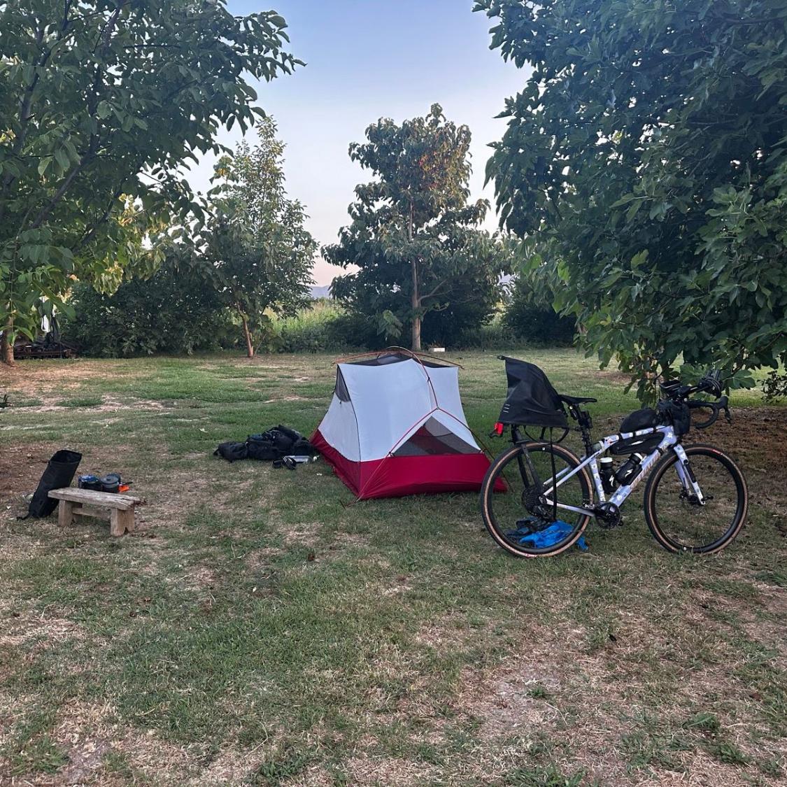 Camping scene with a tent, bicycle, and backpack in a grassy area with trees.
