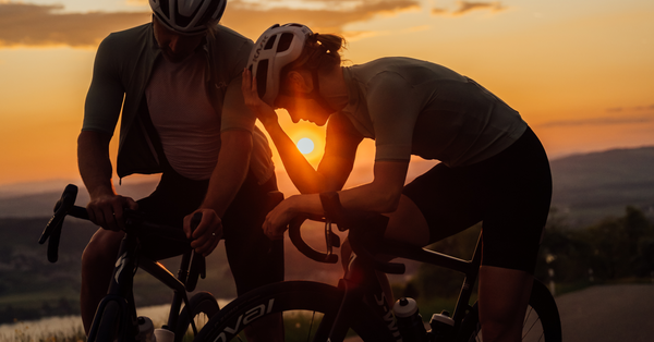Male and female cyclist resting on bike with the setting sun shining between them 